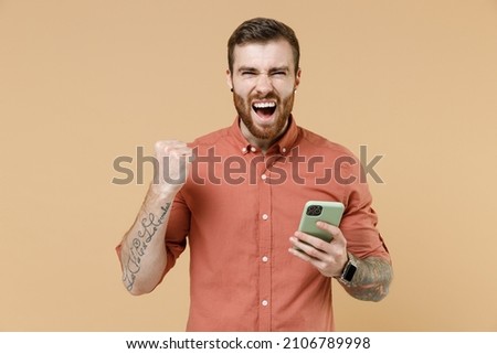 Fun tatooed young brunet man 20s short haircut with earrings wear apricot shirt hold in hand use mobile cell phone doing winner gesture celebrating isolated on pastel orange background studio portrait