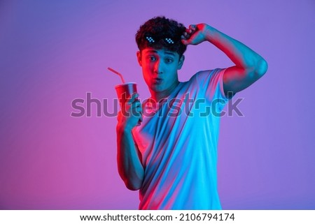 At cinema, movie. Stylish young asian man isolated over lilac color studio background in pink neon light. Concept of human emotions, facial expression, youth culture, diversity