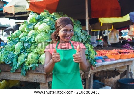 Happy African business woman or female trader wearing a green apron, doing thumbs up gestures while standing at her stall of vegetables in a market place Royalty-Free Stock Photo #2106888437