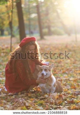 A girl with long red hair, in a red coat and beret, gently strokes her corgi dog in an autumn park. Golden autumn, walking the dog, friendship. beautiful autumn picture