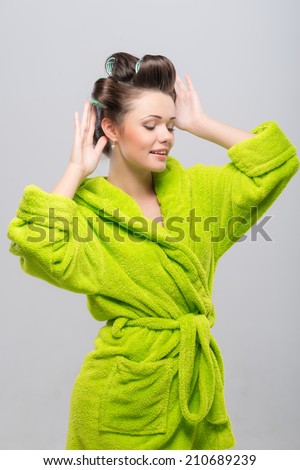 Half-length portrait of pleasant young housewife wearing curlers and green wrapper touching her hair with closed eyes. Isolated on blue background