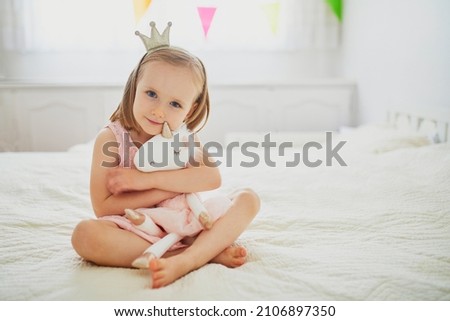 Adorable little girl in pink dress and golden crown dressed as princess playing with unicorn. Kid having fun with soft toy. Child playing with stuffed toy Royalty-Free Stock Photo #2106897350