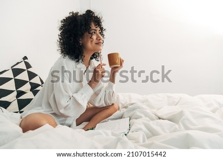 Young mixed race woman sitting on bed having coffee and writing on her journal. Copy space. Royalty-Free Stock Photo #2107015442