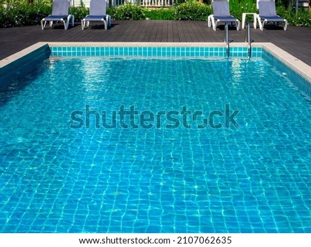 Swimming pool with clean clear water, nobody. Square shaped pool with blue tiles with grab bars ladder and sunbeds on the wood decks, no people. water surface. Overhead view.  Summer background.