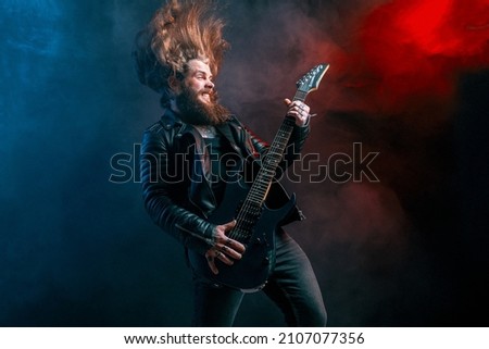 Emotional expression rock guitar player with long hair and beard plays on the black background. Smoke background. Studio shot.  Royalty-Free Stock Photo #2107077356