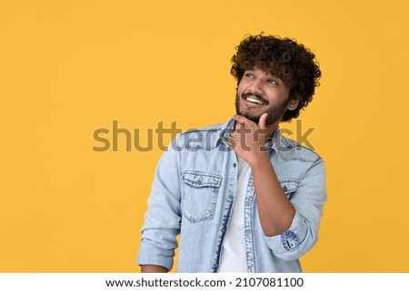 Smiling curious young indian man holding hand on chin looking interested aside at copy space isolated on yellow background thinking of shopping opportunities, planning purchase or dreaming concept. Royalty-Free Stock Photo #2107081100