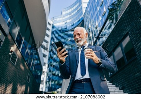 An old executive reading message on the phone at the business center. Low angle view of a happy senior businessman standing at the business center and reading a message on the phone. Royalty-Free Stock Photo #2107086002