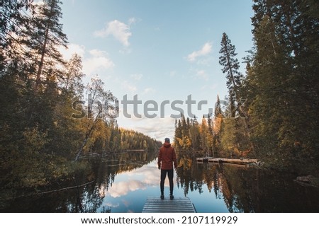 Tireless traveller stands at the end of the pier, admiring the beauty of a stunning autumn in the desolate wilderness of Hossa National Park in Lapland, northern Finland. Royalty-Free Stock Photo #2107119929