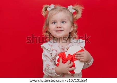 A little cute adorable happy baby kid girl with blond hair in dress holds a red heart shaped gift box in her hands, isolated on red background. Valentine's day shopping concept. Christmas time