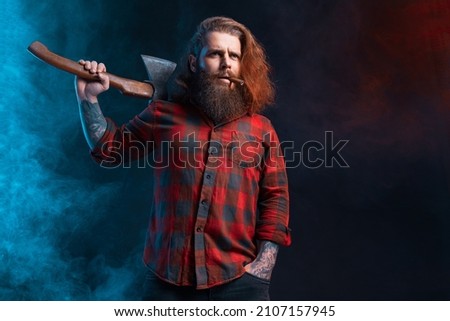 Bearded man with cigar holds old axe. Bearded lumberjack. Serious brutal male in checkered shirt with tattoos with long hair. Studio shot. Smoke background  Royalty-Free Stock Photo #2107157945