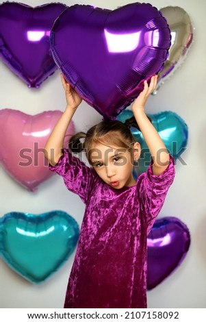 A girl holds a purple heart-shaped balloon above her head, smiling sweetly. Background with decorations from foil balloons for Valentine's day