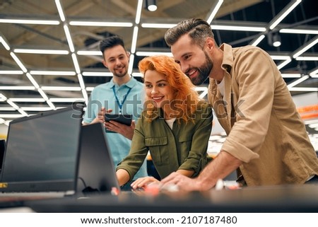 Young couple receives advice from seller in store of household appliances and gadgets, buying new laptop. Male consultant helps in choosing laptop. Royalty-Free Stock Photo #2107187480