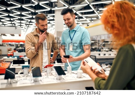 Sales consultant advises man who chooses new smartphone in store of household appliances, electronics and gadgets. Professional consultation in store. Sale Day. Royalty-Free Stock Photo #2107187642