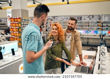 Young couple receives advice from seller in store of household appliances and gadgets, buying new laptop. Male consultant helps in choosing laptop.