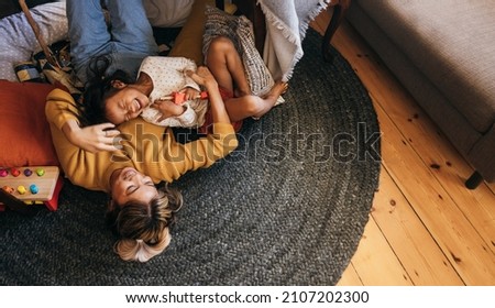 Overhead view of a mother and her daughter laughing happily at home. Cheerful mother and daughter lying on the floor in their play area. Mother and daughter spending some quality time together. Royalty-Free Stock Photo #2107202300