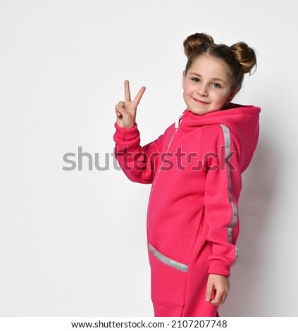 cool girl in a pink jumpsuit with a hood and reflective safety stripes is standing with her back to the camera and turned around showing a victory sign with her hand on a light background