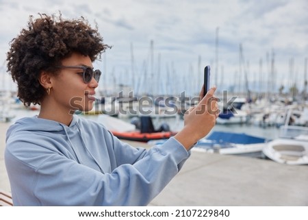 Sideways shot of happy curly haired woman takes photo on mobile phone of harbor view wears sunglasses and sweatshirt enjoys good rest uses modern gadget speds free time at touristic sea resort