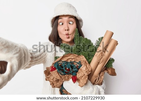 Funny brunette young woman makes fish lips crosses eyes foolishes around shows grimace keeps arm outstretched for making selfie wears white panama warm knitted sweater prepares for New Year. Royalty-Free Stock Photo #2107229930