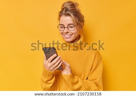 Modern woman with combed hair holds mobile phone reads screen or chats glad to receive pleasant message wears round spectacles casual jumper isolated over yellow background downloads news application Royalty-Free Stock Photo #2107230158