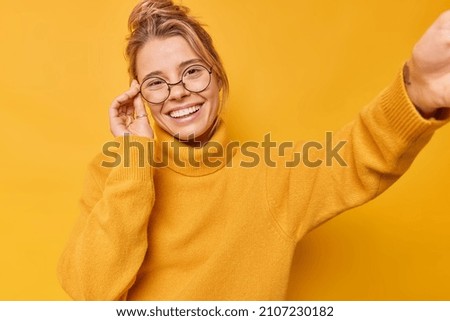 Attrctive woman had delightful look smiles happily stretches arm to camera poses for making selfie wears round eyeglasses and sweater isolated over yellow background. Striking pose for photo Royalty-Free Stock Photo #2107230182