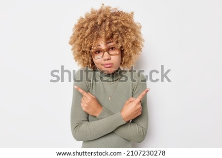 Hesitant clueless woman with curly hair points at different sides picks two variants hesitates about something chooses between wears eyeglasses and turtleneck isolated over white background. Royalty-Free Stock Photo #2107230278