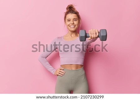 Sporty young woman being in good physical shape keeps hand on waist raises dumbbell has regular fitness training wears sportsclothes poses against pink background. Gym workout and sport concept Royalty-Free Stock Photo #2107230299
