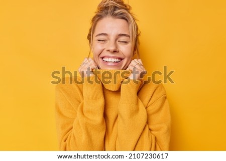 Happy optimistic woman keeps eyes closed smiles gently daydreams holds hands on collar of jumper recalls nice memories isolated over vivid yellow background breathes free. Carefree emotions. Royalty-Free Stock Photo #2107230617