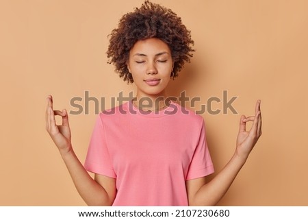 Calm concentrated young woman keeps eyes closed fingers together meditates indoor practices yoga wears casual pink t shirt isolated over brown background tries to find peace stands in zen pose