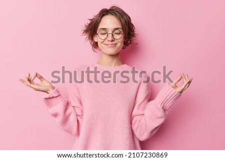 Waist up shot of relaxed happy young woman makes zen gesture meditates indoor breathes deeply practices yoga to feel relaxed wears spectacles and casual jumper isolated over pink background. Royalty-Free Stock Photo #2107230869