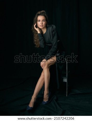 Seductive young woman model with long curly hair and bare legs in elegant cocktail dress sits on bar stool on black background in studio Royalty-Free Stock Photo #2107242206
