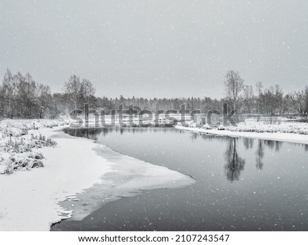 Snowfall on the river. Winter landscape with small river in wood on background gray snowy sky.