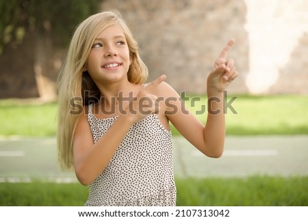 beautiful Caucasian little kid girl wearing dress standing outdoors smile excited directing fingers look empty space