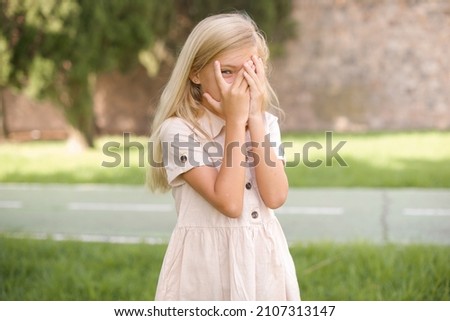 beautiful Caucasian little kid girl wearing dress standing outdoors covering face with hands and peering out with one eye between fingers. Scared from something or someone.