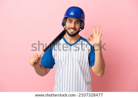 Baseball player with helmet and bat isolated on pink background showing ok sign with fingers