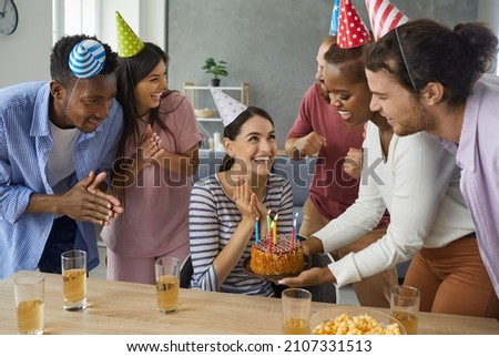 Diverse group of cheerful friends in their twenties giving a cake with burning candles to a happy birthday girl. Young woman in her 20s getting presents and having fun at her birthday party at home