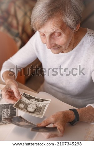 Mature woman watching black and white old photo album at home. Grandmother is looking at her own photo was taken in 1957 She has got smile while remembering how young she was. Selective focus.