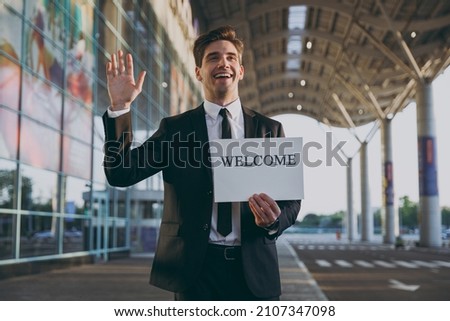 Bottom view young satisfied traveler businessman man in black suit stand outside at international airport terminal hold card sign with welcome title text waving hand Air flight business trip concept.