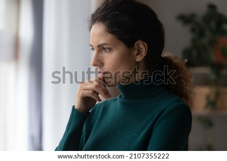 Thoughtful unhappy jealous young hispanic latin woman touching chin, standing near window, considering problem solution or feeling stressed of difficult life situation, bad mood depression concept. Royalty-Free Stock Photo #2107355222