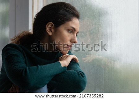 Thoughtful stressed young hispanic latin woman sitting on windowsill, looking outside on rainy weather, having depressive or melancholic mood, suffering from negative thoughts alone at home. Royalty-Free Stock Photo #2107355282
