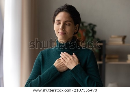 Head shot peaceful young attractive woman holding folded hands on chest, praying waiting for miracle, feeling thankful indoors. Sincere happy female volunteer showing kindness or expressing gratitude. Royalty-Free Stock Photo #2107355498