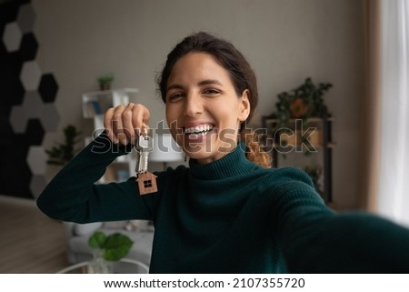 Happy young woman holding keys in hands, recording selfie video, sharing positive experience of apartment purchase, renting first dwelling or moving into renovated house, blogging streaming online.