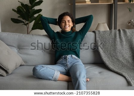 Happy young hispanic woman sitting on comfortable couch, feeling refreshed after day dream, meditating alone in living room. Mindful millennial lady relaxing with folded hands behind head at home. Royalty-Free Stock Photo #2107355828