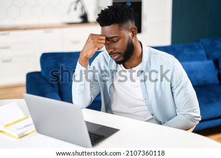 Tired frustrated man sitting with eyes closed and using laptop at home, holding bridge of the nose, feels despair, African-American male student fail exams, has problems with remote work