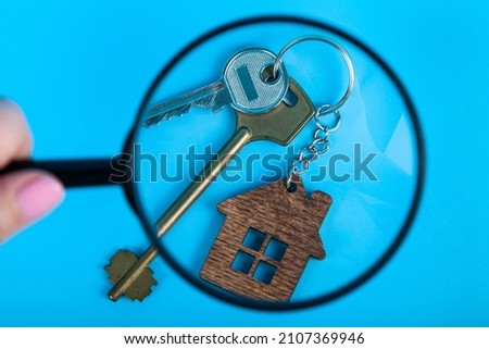 Keys with a keychain in the form of a house under magnifying glass. Rent apartments, Real Estate and buying a house idea. Blue background