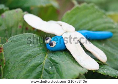Plasticine world - little homemade blue dragonfly with white wings sitting on a green leaf, selective focus on head