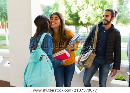Happy young woman giving a kiss on the cheek to her best friend at the university while carrying books to class Royalty-Free Stock Photo #2107398077