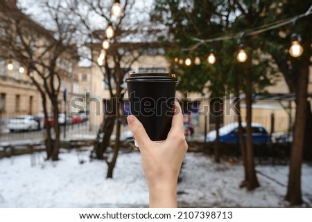 A paper cup with coffee, cocoa or tea in the hands of a girl against the backdrop of the atmosphere of the Christmas city. Time to drink hot holiday drinks. Empty space for text, layout. Coffee to go