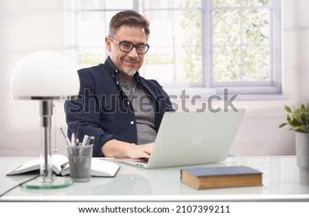 Casual businessman working with laptop computer in home office, happy, smiling. Portrait of middle age man in 50s, white caucasian, gray hair. Royalty-Free Stock Photo #2107399211