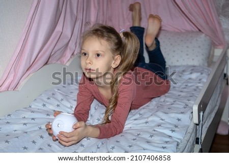 The girl is lying in bed and watching something interesting through the projector.A girl's nursery in pink tones.The girl has a small portable projector in her hands for watching movies and cartoons