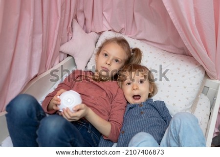 A boy and a girl are lying on the bed and watching cartoons on the ceiling through a projector.A girl is holding a children's mini-projector in the shape of a cube.An attentive and close look